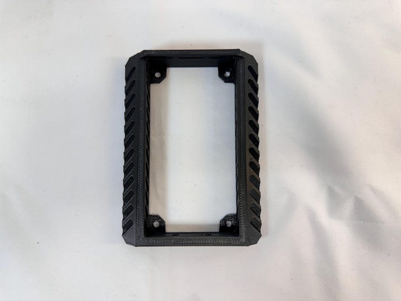 K&ouml;nnte beinhalten: Ein schwarzer rechteckiger Rahmen mit einem strukturierten, geriffelten Design an den Seiten. Der Rahmen hat eine rechteckige &Ouml;ffnung in der Mitte und Befestigungsl&ouml;cher an jeder Ecke. Das Objekt ist wahrscheinlich eine Montagehalterung oder ein Geh&auml;use.