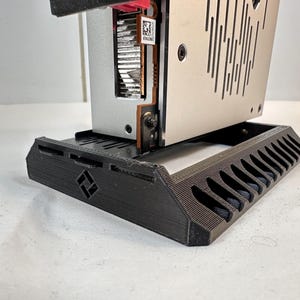 K&ouml;nnte beinhalten: Ein schwarzer, 3D-gedruckter Computerkomponentenst&auml;nder mit geometrischem Design. Der St&auml;nder tr&auml;gt eine silberne und schwarze Computerkomponente mit sichtbarem rotem Licht. Der St&auml;nder hat eine Reihe von abgewinkelten Schlitzen.