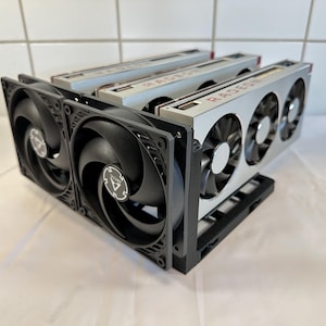 Puede incluir: Tres tarjetas gráficas Radeon plateadas dispuestas en fila paralela, cada una con un sistema de refrigeración de tres ventiladores. Las tarjetas están montadas en un marco de plástico negro con dos ventiladores Arctic negros adicionales al frente.