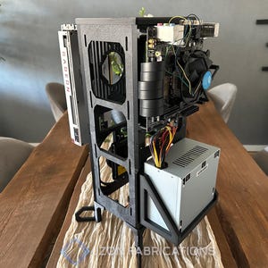 May include: A vertical open-frame computer chassis constructed from black material, featuring a mounted Radeon graphics card, a motherboard with a cooling fan, and a silver power supply unit. The frame includes structural cutouts and wiring.