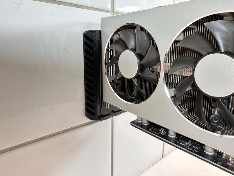 K&ouml;nnte beinhalten: Eine silberne und schwarze Computer-Grafikkarte mit zwei gro&szlig;en L&uuml;ftern. Die L&uuml;fter haben schwarze Fl&uuml;gel und sind in einem silbernen Geh&auml;use untergebracht. Eine schwarze Halterung ist zur Montage an der Seite angebracht.