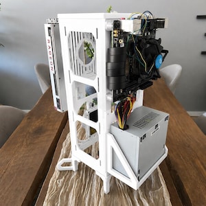 May include: A white open-frame computer case featuring a vertical RADEON graphics card, a motherboard with a black cooling fan, and a silver power supply unit. The structure is built with white panels and includes visible internal wiring.