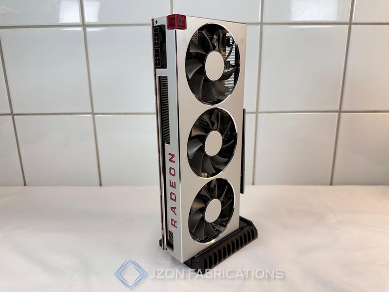 K&ouml;nnte beinhalten: Silberne und schwarze Radeon-Grafikkarte mit drei L&uuml;ftern. Das Wort "RADEON" ist rot auf der Seite aufgedruckt. Die Karte ist auf einem schwarzen St&auml;nder montiert.