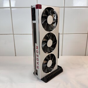 K&ouml;nnte beinhalten: Eine silberne und schwarze Radeon-Grafikkarte mit drei gro&szlig;en L&uuml;ftern. Das Wort "RADEON" ist vertikal in Rot auf der Seite aufgedruckt. Die Karte ist auf einem schwarzen St&auml;nder montiert.