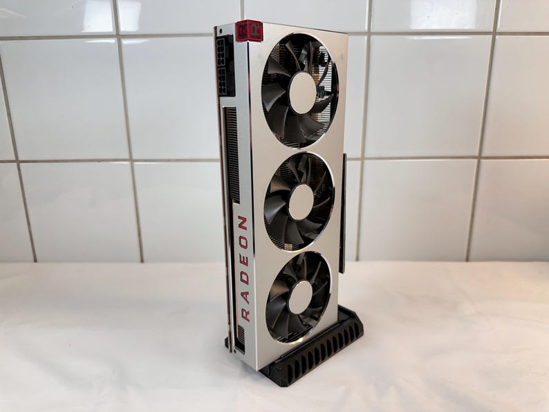 K&ouml;nnte beinhalten: Eine silberne und schwarze Radeon-Grafikkarte mit drei gro&szlig;en L&uuml;ftern. Das Wort "RADEON" ist vertikal in Rot auf der Seite aufgedruckt. Die Karte ist auf einem schwarzen St&auml;nder montiert.