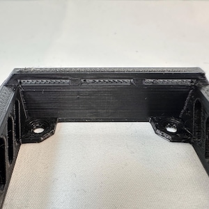 K&ouml;nnte beinhalten: Eine schwarze, rechteckige, 3D-gedruckte Halterung mit rechteckigen Aussparungen an den Seiten. Die Halterung hat zwei kreisf&ouml;rmige L&ouml;cher an den unteren Ecken zur Befestigung. Die Oberkante hat kleine, horizontale Schlitze.