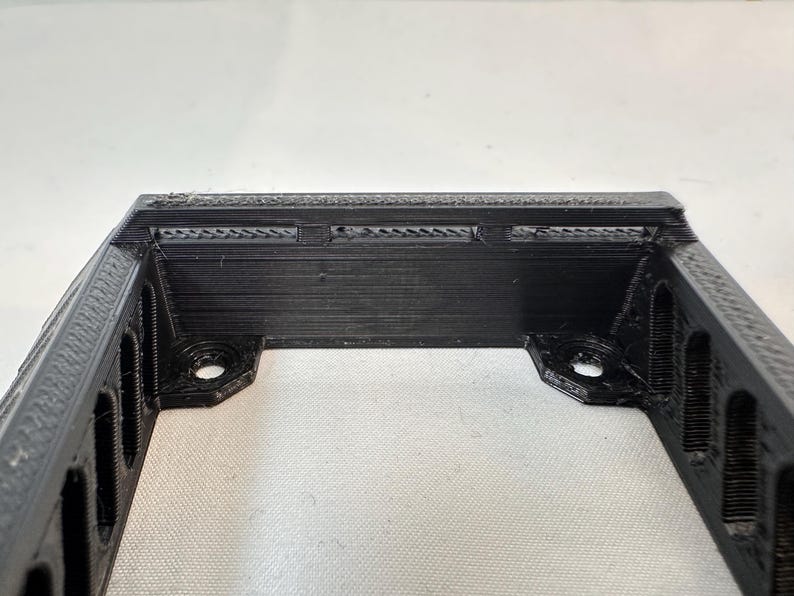 K&ouml;nnte beinhalten: Eine schwarze, rechteckige, 3D-gedruckte Halterung mit rechteckigen Aussparungen an den Seiten. Die Halterung hat zwei kreisf&ouml;rmige L&ouml;cher an den unteren Ecken zur Befestigung. Die Oberkante hat kleine, horizontale Schlitze.
