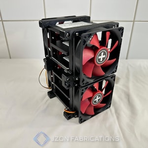Puede incluir: Un conjunto de ventiladores de refrigeración de doble pila con dos marcos cuadrados negros y aspas rojas. El centro de cada ventilador muestra un logotipo de signo más blanco. La unidad tiene soportes negros y cables visibles.