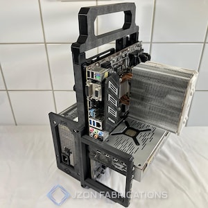 May include: A black open-frame computer chassis with a handle, featuring a motherboard with various ports, a large silver metal CPU cooler, and a power supply unit. The frame is labelled JZON FABRICATIONS at the base, showcasing a modular design.