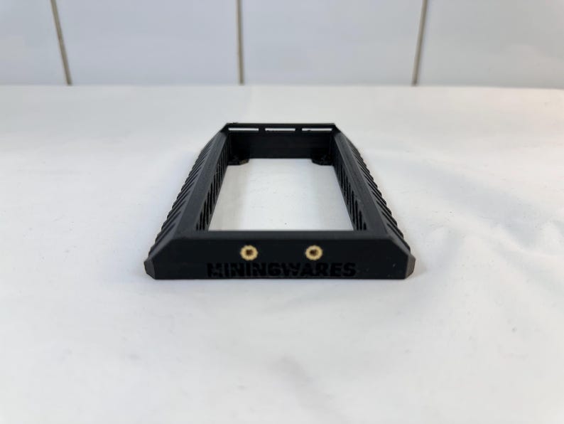 K&ouml;nnte beinhalten: Ein schwarzer rechteckiger Rahmen mit geriffeltem Design an den Seiten. Zwei kleine, goldfarbene kreisf&ouml;rmige Merkmale sind unten sichtbar, mit dem Wort "MININGWARES" darunter gedruckt. Der Rahmen besteht aus mattem schwarzem Material.