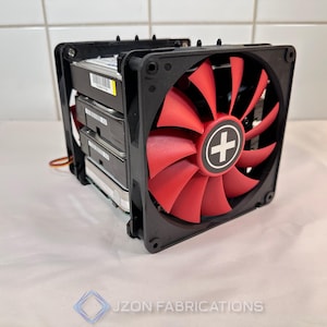 May include: A black computer fan with red blades and a white cross in the center, mounted on a black frame. The fan is positioned in front of a stack of hard drives. The image is on a white surface.