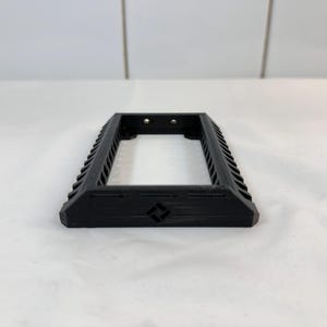 K&ouml;nnte beinhalten: Ein schwarzer rechteckiger Rahmen mit gerilltem Design an den Seiten. Der Rahmen hat eine offene Mitte und zwei kleine L&ouml;cher oben. Ein kleines geometrisches Symbol ist auf der Vorderseite des Rahmens sichtbar.