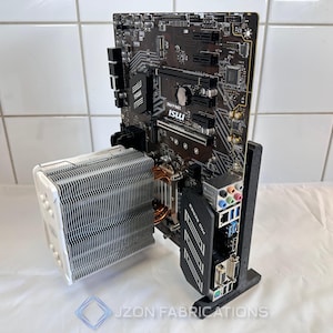 Puede incluir: Una placa base negra MSI PRO SERIES con un enfriador de CPU plateado y blanco está montada en un soporte negro. La placa base tiene varios puertos y ranuras, y el enfriador tiene una gran pila de aletas. El soporte tiene el texto "JZON FABRICATIONS".
