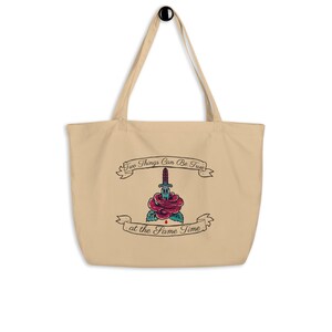 Può includere: Borsa tote beige con manico nero e grafica in stile tatuaggio. La grafica presenta una rosa rossa trafitta da un pugnale, con striscioni sopra e sotto che recitano "Two Things Can Be True" e "at the Same Time."