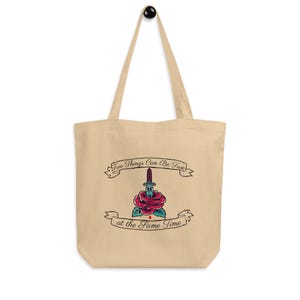 Può includere: Borsa tote beige con manico nero. Presenta una grafica di una rosa trafitta da un pugnale, con il testo "Two Things Can Be True" sopra e "at the Same Time" sotto.