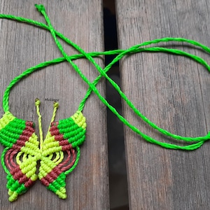 Puede incluir: Un collar con colgante de mariposa de macramé hecho a mano. El diseño de la mariposa presenta colores verde, amarillo y granate. El cordón del collar es de color verde brillante. El collar se exhibe sobre una superficie de madera.
