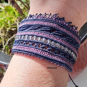 May include: A handmade macrame bracelet in shades of navy blue and pink, adorned with clear beads. The bracelet features intricate knotting and a delicate fringe detail along the top edge. The word "Meliame" is visible on the bracelet.