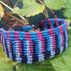 May include: A macrame bracelet in a woven pattern, featuring a combination of pink, blue, black, and white threads. The bracelet is displayed against a backdrop of green leaves.