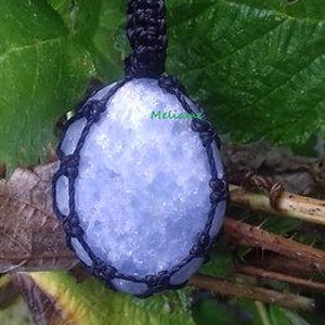 May include: A light blue crystal pendant encased in a black macrame net. The pendant is oval-shaped and has a textured surface. The macrame cord forms a loop at the top for hanging. The background features green leaves and brown twigs.
