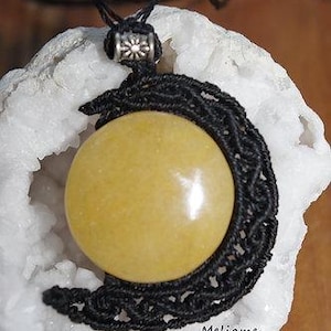 May include: A handcrafted pendant featuring a large, round, yellow gemstone set within a black macrame crescent moon design. The pendant hangs from a black cord and is accented with a silver-colored bead. The word "Meliame" is visible.