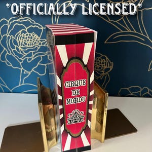 May include: A book-shaped item with a red and white striped top and the words "CIRQUE DE MORDU" on the front. The item is held by two gold-colored bookends. The text "OFFICIALLY LICENSED" is at the top.