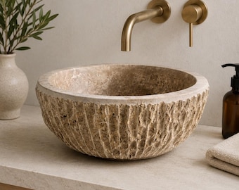 Puede incluir: Un lavabo de sobreponer redondo fabricado en piedra travertino beige natural, con una superficie exterior texturizada y cincelada y un interior liso. El lavabo se combina con un grifo de pared de latón cepillado y un mando monomando a juego.