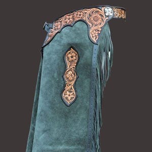 May include: Green suede chaps with intricate floral tooled leather accents. The chaps feature a fringed hem and a decorative leather waistband. The leather detailing includes a floral design. A stylish piece for Western wear.