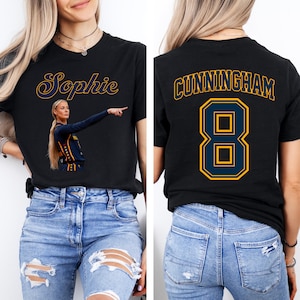 Puede incluir: Una camiseta negra con un gráfico de una jugadora de baloncesto señalando con el nombre Sophie impreso encima en amarillo y azul. La parte posterior de la camiseta muestra el nombre CUNNINGHAM en texto amarillo y azul sobre el gran número 8.