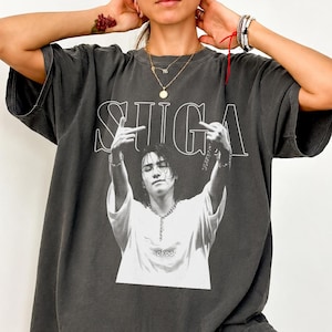 Op de afbeelding: Een antracietgrijs t-shirt met korte mouwen en een grote witte grafische print van een persoon met lang haar die een ketting draagt en handgebaren maakt. De tekst SUGA staat in grote witte omtrekletters boven de persoon gedrukt.