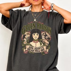 May include: A charcoal gray short-sleeve t-shirt featuring a graphic print with the text THE GREEN RIBBON AND JENNY'S HEAD FELL OFF. The illustration depicts a central figure with a green ribbon around their neck, surrounded by smaller scenes.