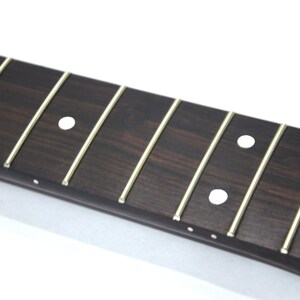 May include: Close-up of a guitar fretboard with dark brown wood and thin, gold-colored frets. White dot inlays mark the fret positions. The wood grain is visible, and the frets are evenly spaced.