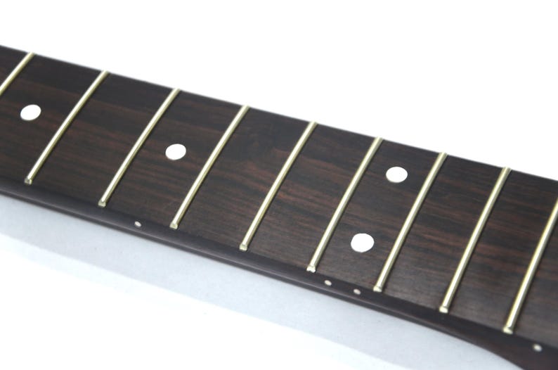 May include: Close-up of a guitar fretboard with dark brown wood and thin, gold-colored frets. White dot inlays mark the fret positions. The wood grain is visible, and the frets are evenly spaced.
