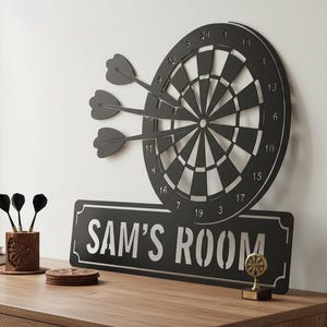 May include: A black metal dartboard with three darts embedded, mounted on a white wall. Below the dartboard is a black sign that reads "SAM'S ROOM" in white lettering. A wooden table holds darts, coasters, and a small trophy.