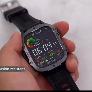 May include: A black smartwatch with a gray bezel and black band displays the time, date, and health metrics. The screen shows "06:04 55" and "100". The watch is held in a hand, and the text "Scratch and abrasion resistant" is visible.