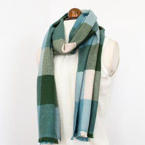May include: A patterned scarf in shades of green, blue, and beige, draped over a mannequin. The scarf features a plaid design with fringed edges. The soft texture and cool colour palette make it a versatile accessory. The scarf is approximately 180 cm long.
