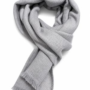 May include: A light grey scarf featuring a dense, textured knit pattern throughout the fabric. The scarf is arranged in a loose loop and finished with short, delicate fringe detailing along the straight edges at both ends of the garment.