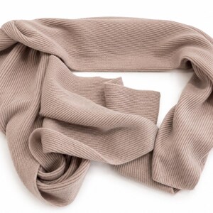 May include: A light taupe scarf featuring a consistent ribbed knit texture throughout the entire length of the fabric. The scarf is arranged in a loose, folded loop shape, showcasing the soft, uniform colour and the vertical lines of the knit pattern.