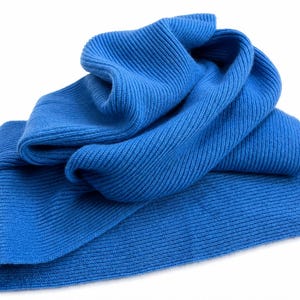 May include: A bright blue scarf featuring a distinct ribbed knit texture. The fabric is folded over itself, creating soft, layered curves and deep ridges throughout the material. The scarf has a consistent, vibrant blue colour across its entire surface.