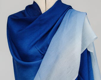 May include: A draped blue shawl with a gradient effect, transitioning from a deep indigo to a light, airy blue. The fabric is soft and appears to be lightweight, perfect for layering. The shawl is displayed on a mannequin.