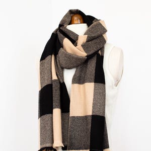 May include: A large scarf with a black, beige, and grey checked pattern. The scarf is draped over a white mannequin. The scarf has a fringed edge and is made of a soft material.