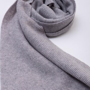 May include: A grey knitted scarf, folded and draped, with a ribbed texture. The scarf is a solid, neutral colour, and appears soft and warm. The fabric is tightly woven, suggesting a high-quality material.