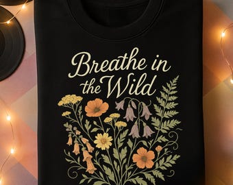 May include: Black sweatshirt with the text "Breathe in the Wild" in a cream-colored script. The design features a floral arrangement with orange, yellow, and purple flowers and green leaves. The sweatshirt is folded.