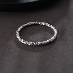 May include: A silver ring featuring a twisted rope pattern. The ring is circular and rests on a dark grey surface. The texture of the ring is detailed, with each twist clearly visible. The overall design is simple and elegant.