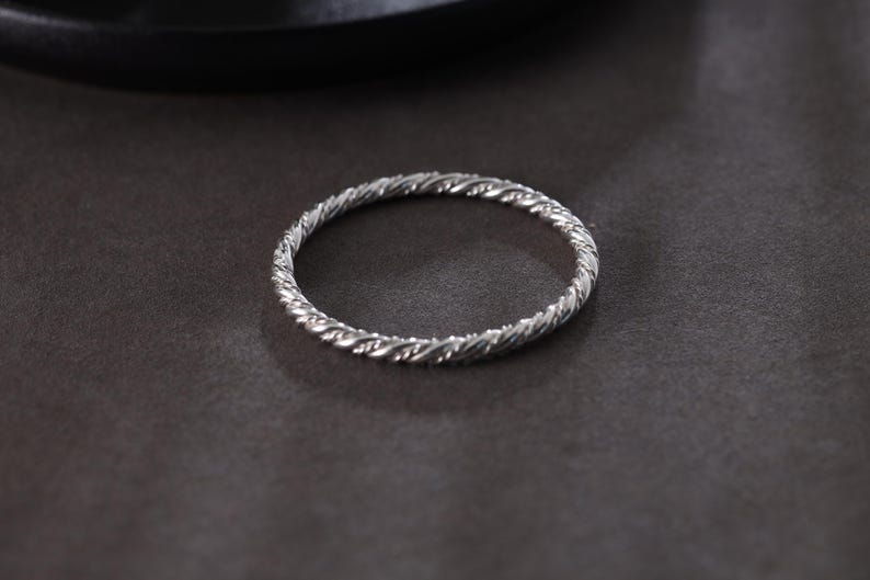 May include: A silver ring featuring a twisted rope pattern. The ring is circular and rests on a dark grey surface. The texture of the ring is detailed, with each twist clearly visible. The overall design is simple and elegant.
