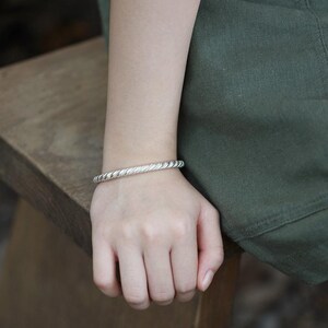 May include: A silver bracelet with a twisted rope design. The bracelet is worn on a wrist, resting on a wooden surface. The person is wearing a white top and green trousers.