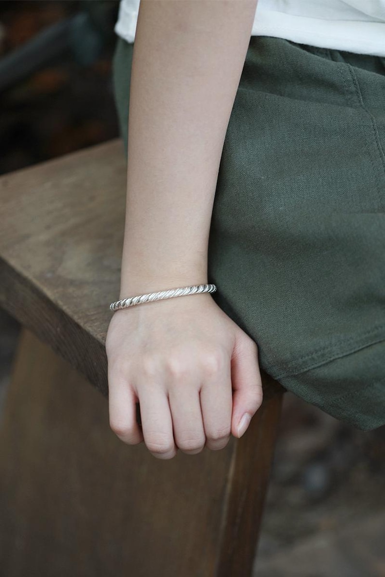 May include: A silver bracelet with a twisted rope design. The bracelet is worn on a wrist, resting on a wooden surface. The person is wearing a white top and green trousers.