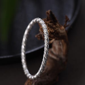 May include: A silver bangle with a twisted rope pattern. The bracelet is shown on a piece of textured, brown wood against a dark backdrop. This piece of jewellery is a simple, elegant accessory.