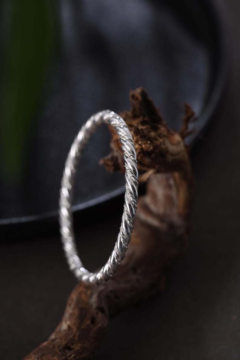 May include: A silver bangle with a twisted rope pattern. The bracelet is shown on a piece of textured, brown wood against a dark backdrop. This piece of jewellery is a simple, elegant accessory.
