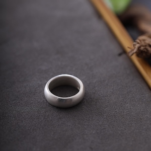 Puede incluir: Un anillo de plata con un diseño liso y redondeado. El anillo está sobre una superficie gris oscuro, con un marco de madera y vegetación en el fondo. El estilo sencillo y elegante del anillo lo hace adecuado para el uso diario.
