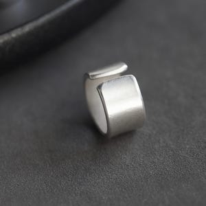 Może przedstawiać: Zbliżenie srebrnego pierścionka o minimalistycznym designie. Pierścionek ma prostokątny kształt z przerwą i jest umieszczony na ciemnoszarym tle. Pierścionek wydaje się być wykonany z metalu.